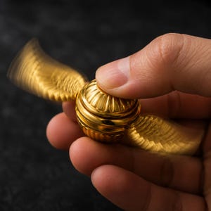 May include: A gold-coloured metal fidget spinner shaped like a sphere with two extended wings. The central spherical component features a ribbed, fluted texture, while the wings appear blurred due to rapid motion, creating a dynamic visual effect.