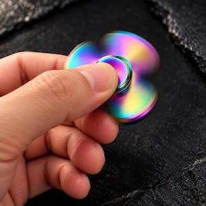 Può includere: Un fidget spinner a tre pale con una finitura metallica arcobaleno. Il dispositivo presenta un tappo centrale circolare e tre bracci arrotondati in movimento, che mostrano una sfumatura di vivaci tonalità viola, blu, verdi e gialle.