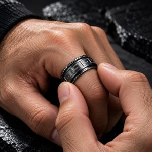 Può includere: Un anello nero caratterizzato da una fascia centrale con un motivo geometrico greco ripetuto. I bordi dell'anello presentano un design testurizzato e dentellato. La superficie del metallo ha una finitura opaca, dall'aspetto scuro e uniforme.