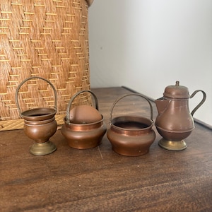 May include: A collection of four miniature copper-toned metal vessels including two small buckets with wire handles, one small bowl with a wire handle containing a brown sphere, and a small lidded pitcher with a handle and a brass-toned base.