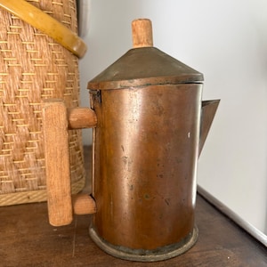 May include: A cylindrical copper coffee pot featuring a flat, conical lid with a small wooden knob handle. The pot has a vertical wooden handle attached to the side and a small, triangular spout. The copper surface shows signs of natural patina.