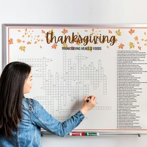 May include: A large white Thanksgiving crossword puzzle with the words "Thanksgiving Meals & Foods" at the top. The crossword is framed and has fall leaf decorations. A person is writing on the crossword with a black marker.