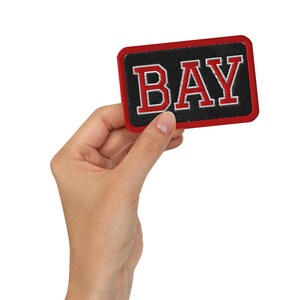 Può includere: Una toppa rettangolare ricamata con sfondo nero e bordo rosso. La parola BAY è cucita in grandi lettere rosse in grassetto con un contorno bianco, caratterizzata da un classico carattere in stile college centrato sulla superficie della toppa.