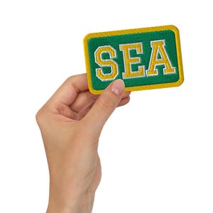 May include: A rectangular patch with the letters "SEA" embroidered in gold on a green background. The patch has a gold border and is held by a hand against a white background.