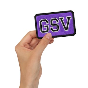 May include: A rectangular purple patch featuring the letters "GSV" in black and white. The patch has a black border and is held by a hand against a white background. The letters are in a bold, collegiate style.