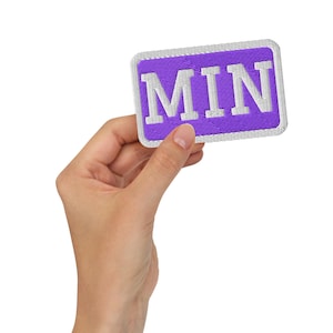 Könnte beinhalten: Ein rechteckiger bestickter Aufnäher mit violettem Hintergrund und weißem Rand. Der Aufnäher zeigt den Text MIN in großen, fetten, weißen Großbuchstaben, die auf dem violetten Stoff zentriert sind. Das Design hat einen sauberen Rand.