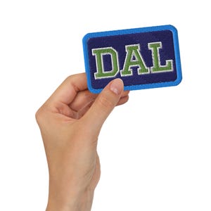 May include: A rectangular patch with the letters "DAL" embroidered in green and white on a navy blue background. The patch has a blue border and is held by a hand.