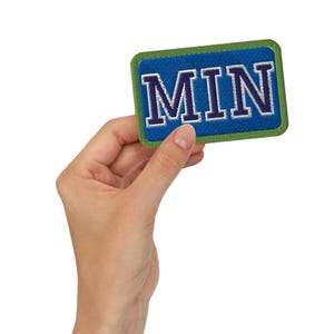 May include: A rectangular patch with the word "MIN" in white, outlined in blue, on a blue background. The patch has a green border. The patch is held in a hand.