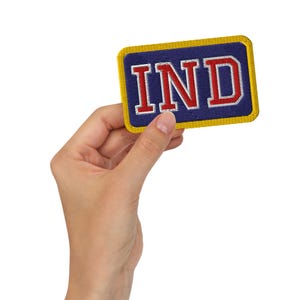 May include: An embroidered patch in a rectangular shape, featuring the letters "IND" in red with a white outline, set against a navy blue backdrop. It has a gold border and is held by a hand.