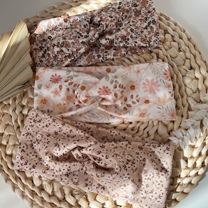 Könnte beinhalten: Drei Haarband-Stirnbänder mit Blumenmuster in Braun-, Rosa- und Cremetönen werden auf einer gewebten Matte präsentiert. Die Stirnbänder weisen verschiedene Blumenmuster auf, darunter kleine und große Blüten und Blätter. Die Stirnbänder sind vorne verdreht.