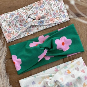 Könnte beinhalten: Drei Stoff-Haarbänder mit gedrehten Knotendetails. Ein Haarband hat einen weißen Hintergrund mit einem Blumenmuster in Rosa, Blau und Grün. Ein weiteres ist grün mit großen rosa Blumen. Das dritte ist weiß mit pastellfarbenen Herzen.