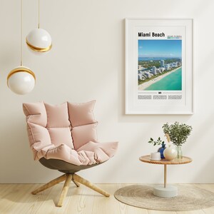 May include: A pink, plush armchair with a wooden base sits in a room with a white wall, a framed poster of Miami Beach, Florida, and a small wooden table with a vase of greenery and a blue ceramic vase.
