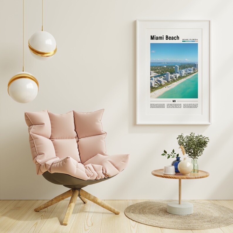 May include: A pink, plush armchair with a wooden base sits in a room with a white wall, a framed poster of Miami Beach, Florida, and a small wooden table with a vase of greenery and a blue ceramic vase.