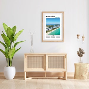 May include: A framed print of an aerial view of Miami Beach, Florida, with the text "Miami Beach" and "South Beach Miami" printed below the image. The print is hanging on a white wall above a light wood cabinet with two doors. A potted plant is on the left side of the cabinet and a small wooden stool with a vase of dried flowers is on the right side of the cabinet.