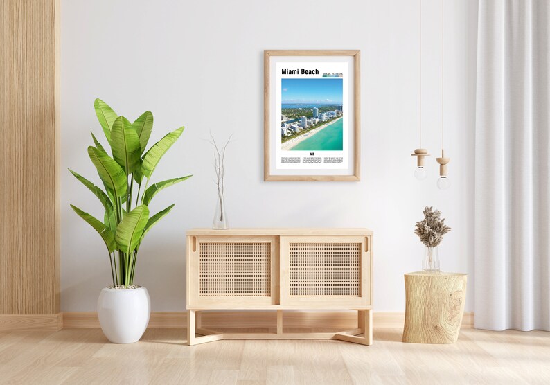 May include: A framed print of an aerial view of Miami Beach, Florida, with the text "Miami Beach" and "South Beach Miami" printed below the image. The print is hanging on a white wall above a light wood cabinet with two doors. A potted plant is on the left side of the cabinet and a small wooden stool with a vase of dried flowers is on the right side of the cabinet.
