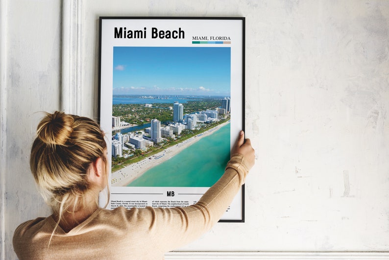 May include: A framed poster with an aerial view of Miami Beach, Florida. The poster has the text "Miami Beach" and "Miami, Florida" at the top. The poster also has the text "MB" at the bottom.