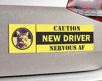 Könnte beinhalten: Ein gelber rechteckiger Autoaufkleber mit dem Text "CAUTION NEW DRIVER NERVOUS AF". Er zeigt auch eine kreisförmige Grafik eines Katzenkopfes. Der Aufkleber ist an der Rückseite eines silbernen Autos angebracht.