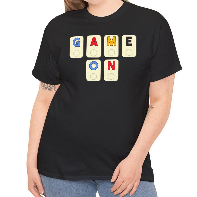 May include: Black t-shirt with the words "GAME ON" spelled out using colorful game tiles. The letters are in blue, red, yellow, and white, with a circular design in the center of each tile. The shirt is a casual style.