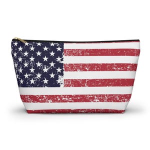 May include: A zippered pouch with a distressed American flag design. The pouch has a black zip and a white interior. The flag design includes red and white stripes and a blue field with white stars.