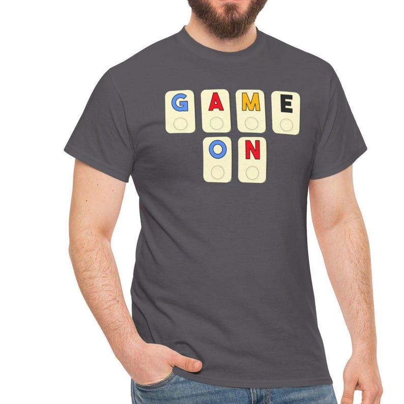 May include: A charcoal gray t-shirt featuring the words "GAME ON" spelled out with colorful letter tiles. The letters are in a playful font, with a blue, red, yellow, and black color scheme. The shirt is a classic crew neck style.