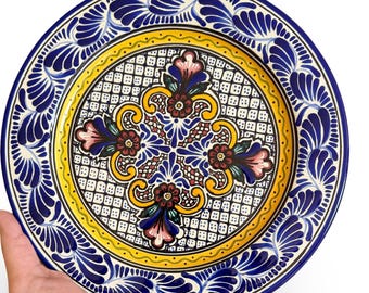 May include: A hand-painted ceramic plate with a vibrant floral design. The plate features a blue border with a repeating leaf pattern, a yellow inner ring, and a central design with blue, pink, and brown floral motifs. The plate is held against a white background.