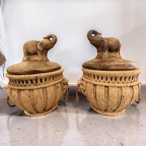 May include: Two decorative ceramic jars with lids. Each jar is beige with a textured surface and a detailed design. The lids are topped with a small elephant sculpture. The jars have handles and are placed on a gray countertop.