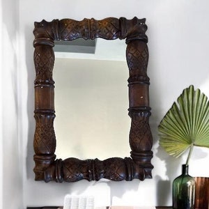 May include: A dark brown, rectangular mirror with ornate carved detailing hangs above a matching wooden cabinet. The cabinet has a woven front and sits on short, angled legs. A green glass bottle and a large green leaf add a touch of nature.