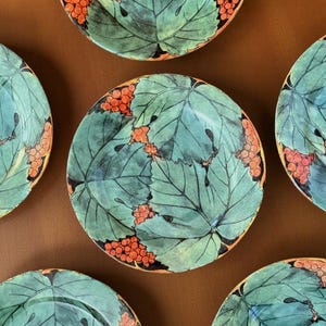 Peut inclure: Six assiettes en céramique avec un motif de feuilles turquoise et de baies oranges. Les assiettes ont une bordure noire et sont disposées sur une surface marron. Les assiettes sont rondes et semblent peintes à la main.