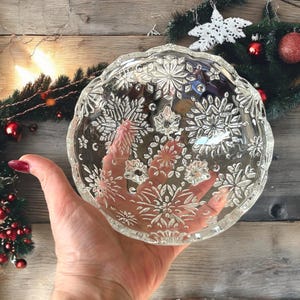 May include: Clear glass bowl with a snowflake design. The bowl has a scalloped edge and is held in a hand. The background includes Christmas decorations with red ornaments and a white star.