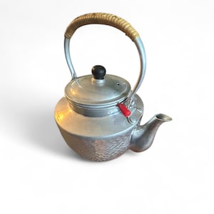 Puede incluir: Una tetera vintage de color plateado con un asa tejida y un pomo negro en la tapa. La tetera tiene un cuerpo texturizado y una palanca roja. El pico es curvo e inclinado.