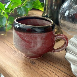 May include: A handmade ceramic mug with a rounded body and a small handle. The mug features a dark red and gray color scheme with a black rim. The mug is sitting on a wooden surface.