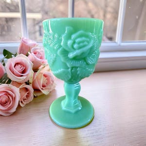 May include: A light green milk glass goblet featuring a raised relief pattern of blooming roses and leaves on the exterior bowl. The glass vessel stands on a pedestal base and is positioned next to a cluster of soft pink roses with green stems.