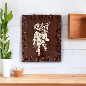 May include: A framed wood carving featuring a figure in a costume with a flowing cape and a mask. The carving is set against a dark brown background within a textured, ornate frame. The artwork is mounted on a white wall.