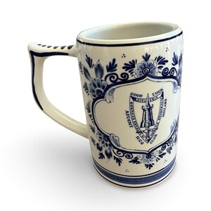 May include: White ceramic mug with a blue floral design and a shield emblem. The mug features a unique handle and a slanted rim. The shield has text that reads "Good Luck" and "Holland".