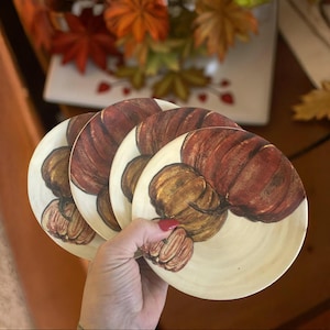 May include: A set of four round coasters with a cream-colored background. Each coaster features a detailed illustration of a pumpkin in shades of brown, orange, and yellow. The coasters are held in a person's hand, with fall foliage in the background.