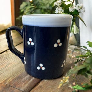 May include: A navy blue ceramic mug with a white rim and handle. The mug is decorated with small white and red floral designs. The mug is sitting on a wooden surface.