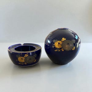 Pode incluir: Um conjunto de dois itens decorativos de cerâmica. Os itens são um cinzeiro azul escuro e um vaso redondo. Ambas as peças apresentam desenhos florais dourados. O cinzeiro tem uma borda dourada. O vaso tem uma pequena abertura.