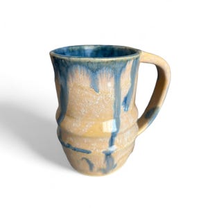 Puede incluir: Taza de cerámica hecha a mano con un diseño único. La taza presenta una base amarilla clara con esmalte azul goteando alrededor del borde y los lados. El asa es de color amarillo claro a juego. Una pieza elegante y funcional para bebidas.