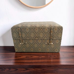 Puede incluir: Una caja de almacenamiento rectangular cubierta de tela con un patrón geométrico repetitivo en verde oliva y dorado con pequeños motivos florales. La tapa se asegura mediante dos correas de tela verticales con cierres de palanca claros.