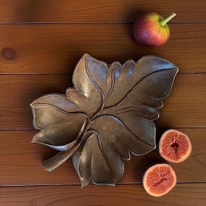 May include: A bronze-colored, leaf-shaped decorative bowl with detailed veining. A red and yellow apple and two halves of a fig are arranged on a wooden surface, complementing the bowl's natural theme. The bowl is a decorative item.