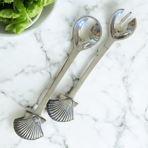May include: A pair of silver salad servers. One server is a spoon, the other a fork. Both have long handles and shell-shaped accents. The servers are on a marble surface, with a bowl of green salad in the background.