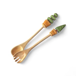 May include: Two wooden salad servers. One is a spoon, the other a fork. The light brown handles are topped with decorative elements: a green cactus and a green leaf. The decorative elements are in small orange pots.