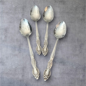 May include: Four vintage silver spoons with detailed floral and scrollwork patterns on the handles. The spoons are arranged on a grey surface, highlighting their polished, reflective surfaces. A classic addition to any dining set.
