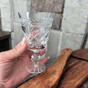 May include: A clear, cut crystal wine glass with a diamond and linear pattern. The glass has a short stem and a round base. The glass is held in a hand, with a wooden cutting board in the background.