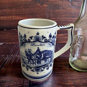 May include: A white ceramic mug with a blue and white design featuring the word "Heineken" and a horse-drawn carriage scene. The mug has a handle and is next to a clear glass bottle.