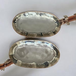 May include: Two oval ceramic serving dishes. The dishes have a light grey interior and a textured, multi-toned border in shades of brown, grey, and blue. The dishes are displayed on a white background.