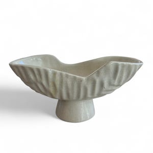 May include: A cream-colored ceramic bowl with a pedestal base. The bowl has a wavy, organic shape with a textured, ridged exterior. The pedestal base is round and supports the bowl. The bowl is likely for decorative or serving purposes.