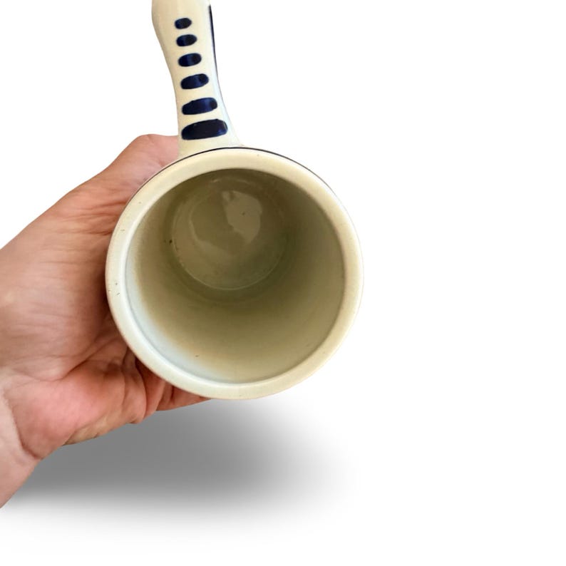 May include: A white ceramic mug with a blue handle and blue accents. The handle has a series of blue oval shapes. The mug is held by a hand, and the interior is a light beige color.