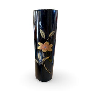 May include: A tall, black vase with a glossy finish. The vase is adorned with a gold-colored floral design featuring a flower and leaves. The flower has a detailed center. The vase is cylindrical and stands upright.