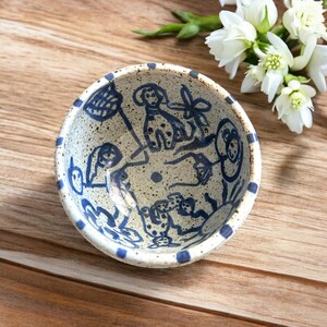 May include: A small, round ceramic bowl with a speckled beige interior and a blue rim. The bowl is decorated with whimsical blue line drawings of dogs and other figures. The bowl is sitting on a wooden surface.